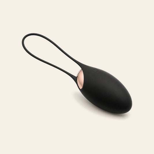 Matte black wireless vibrating egg with rose-gold accent panel and retrieval cord on a soft beige background — Femme Bliss BlissLuna